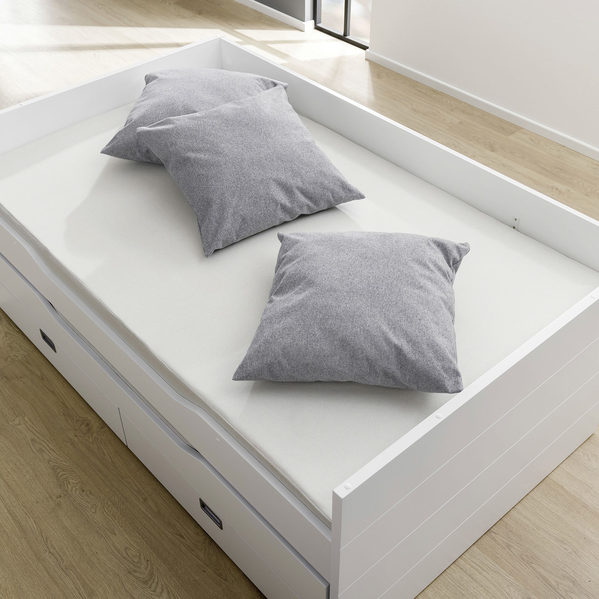 Partykissen 60 x 60 cm, Mikrofaser Steingrau Ein weißes Bett mit Schubladen und drei grauen Kissen auf einer hellen Matratze.