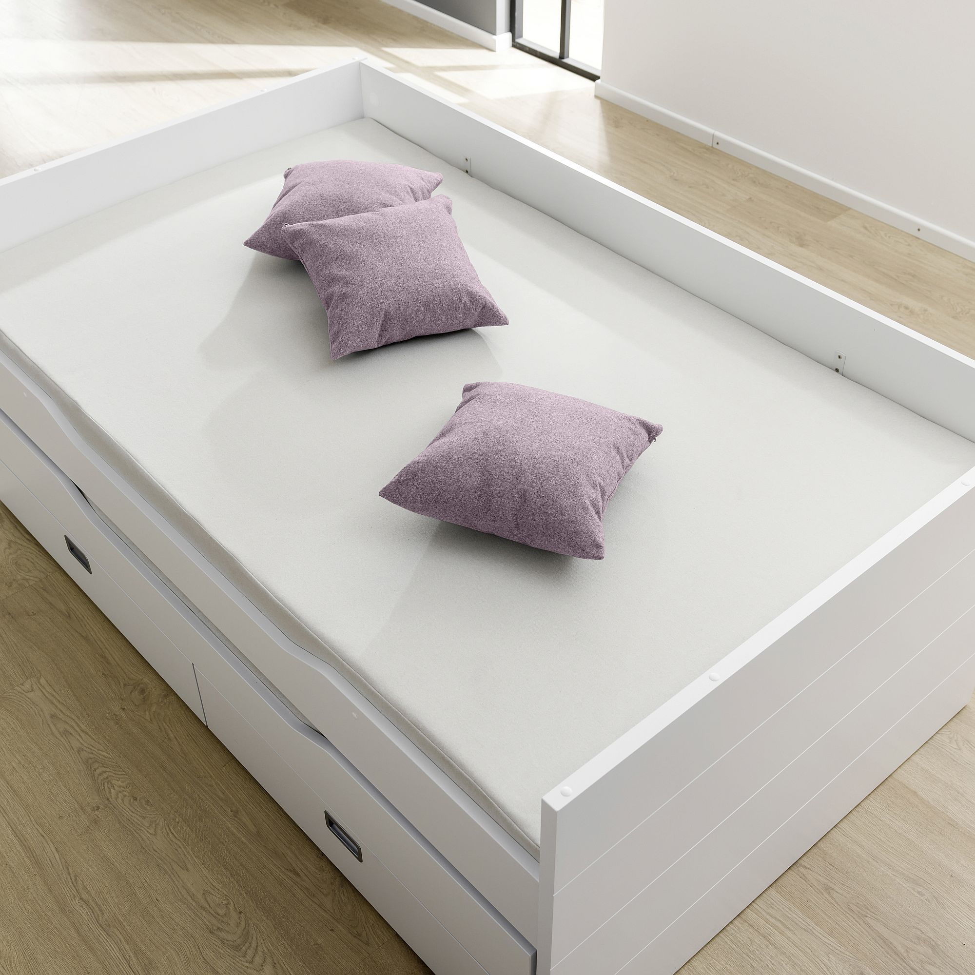 Weißes Bett mit drei hellvioletten Kissen auf einem hellen Holzboden.