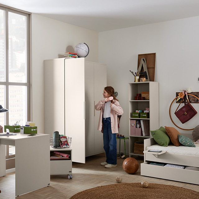 Ein Mädchen steht neben einem weißen Kleiderschrank in einem aufgeräumten Schlafzimmer mit Schreibtisch, Regalen, Sofa und verschiedenen Arbeitsmaterialien.