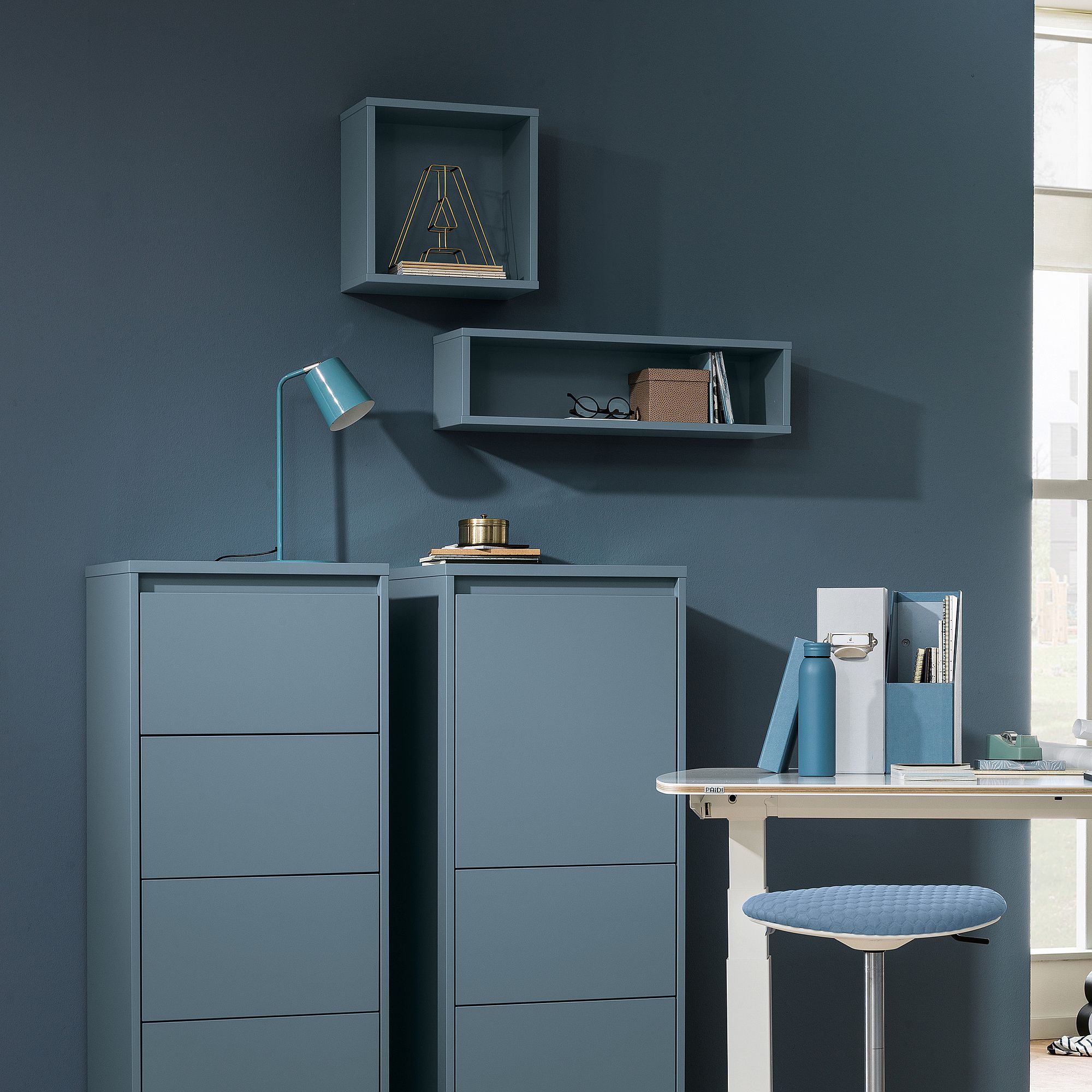Modernes blaues Büro mit Regalen, Schubladen, Lampe und Büchern vor blauer Wand.