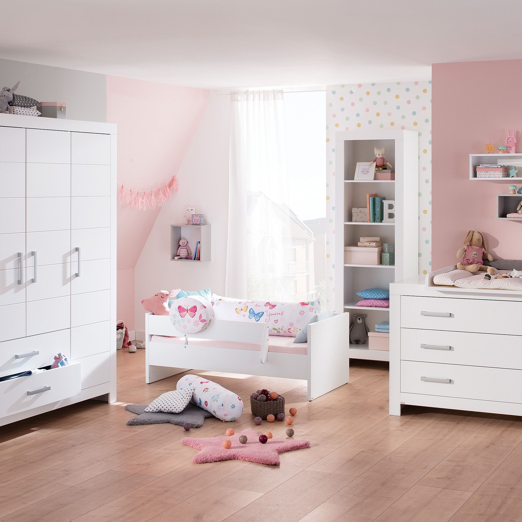 Helles Kinderzimmer mit weißen Möbeln, rosa Akzenten und Spielzeug auf dem Boden.