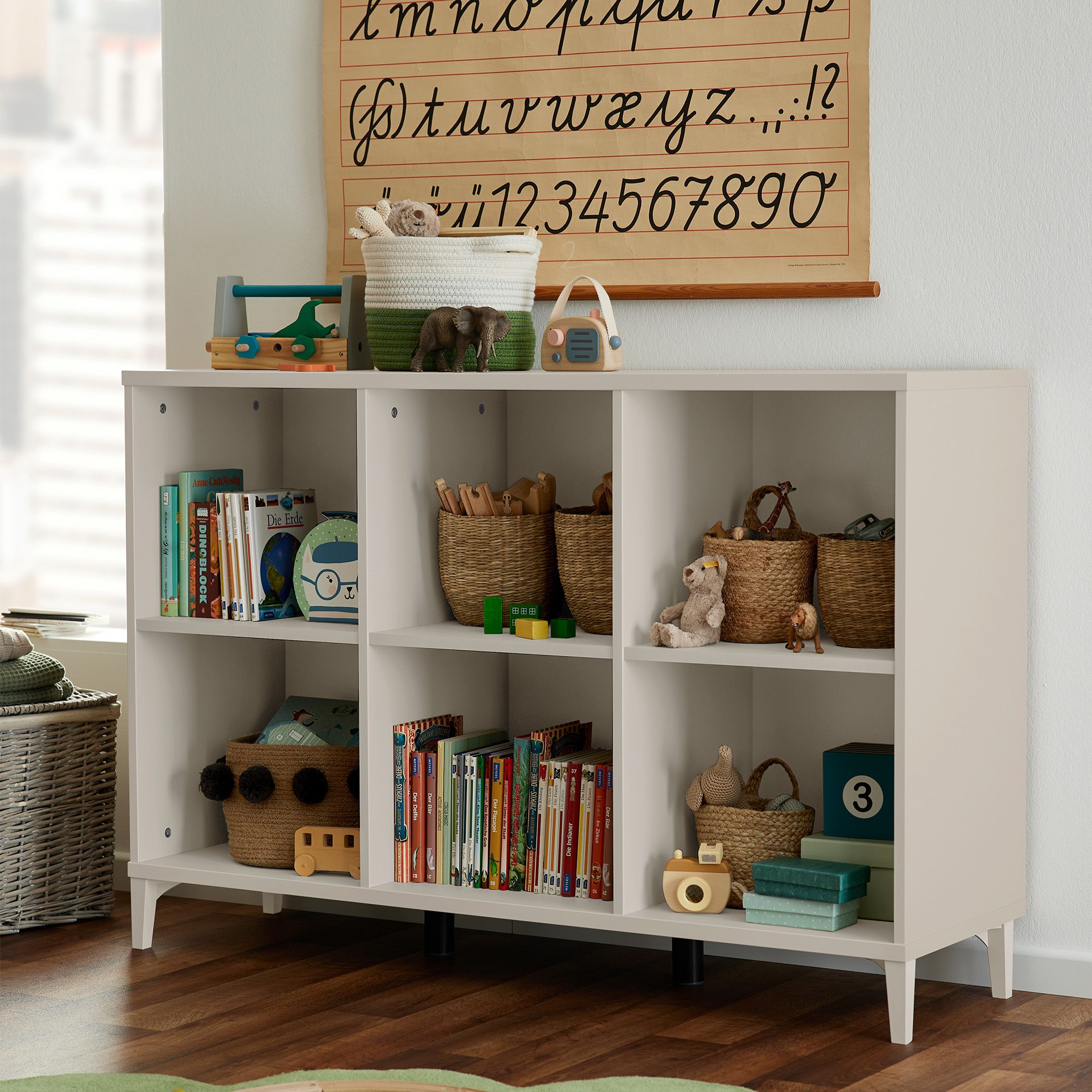 Weißes Regal mit Körben, Spielzeug und Büchern in einem Kinderspielzimmer, darüber eine Buchstabentafel.