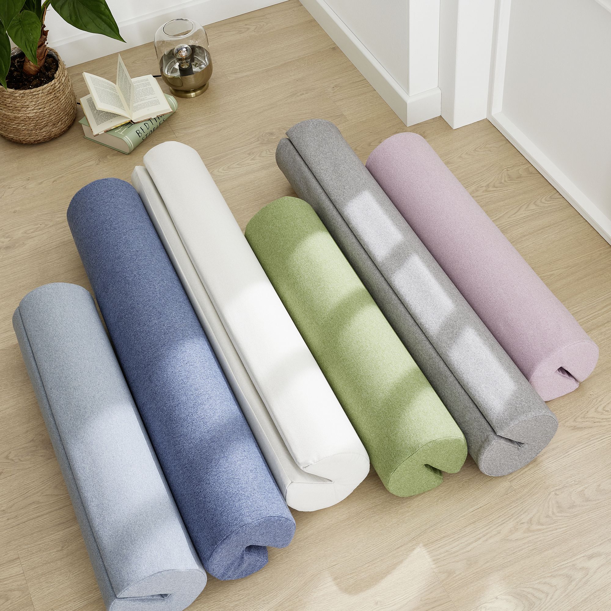 Comfort-Rolle 115 x Ø 20 cm, Mikrofaser Steingrau Sechs gerollte Yogamatten in Pastellfarben auf einem Holzboden neben einem aufgeschlagenen Buch.
