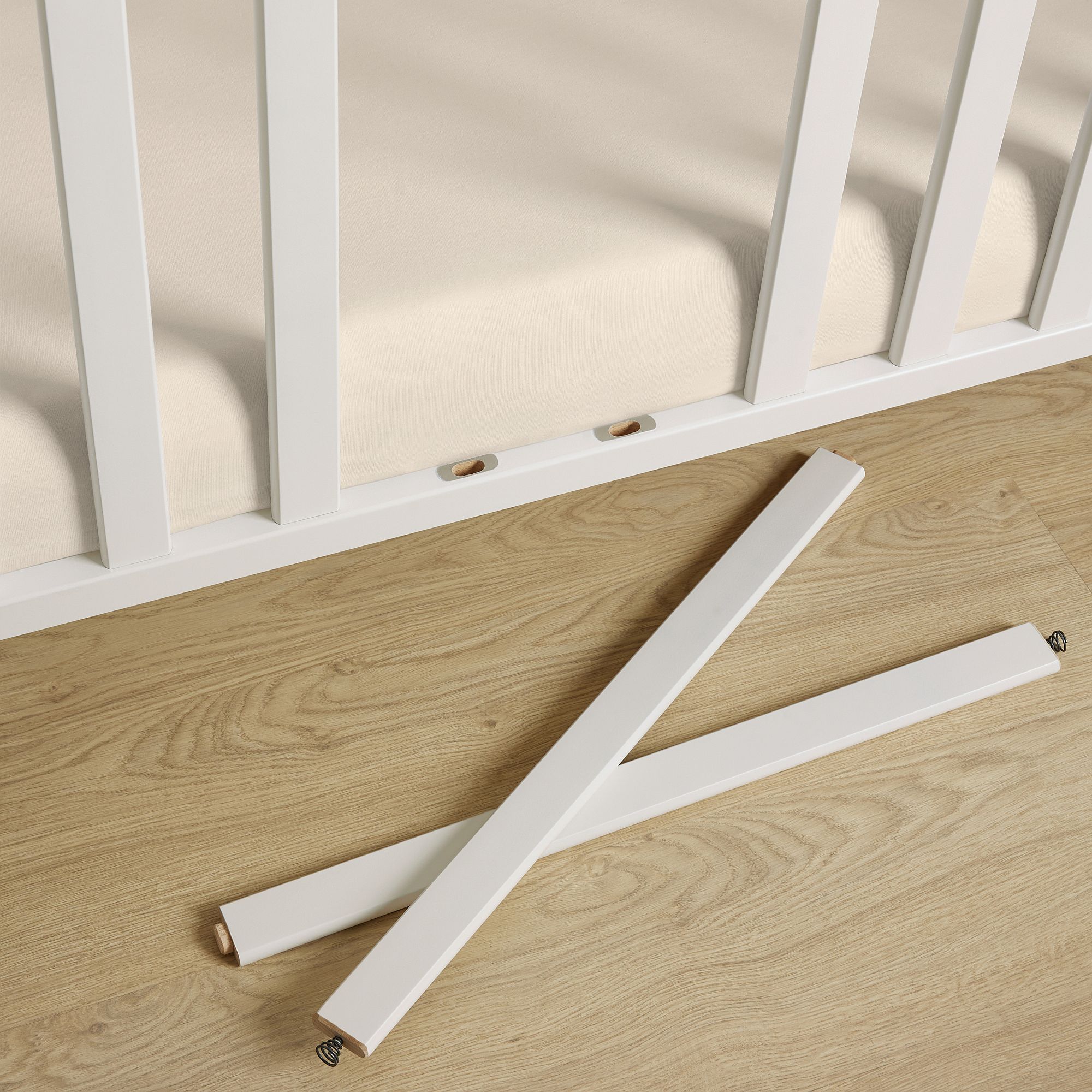 Zwei weiße Krippenstangen liegen gekreuzt auf einem Holzboden vor einem Kinderbett.