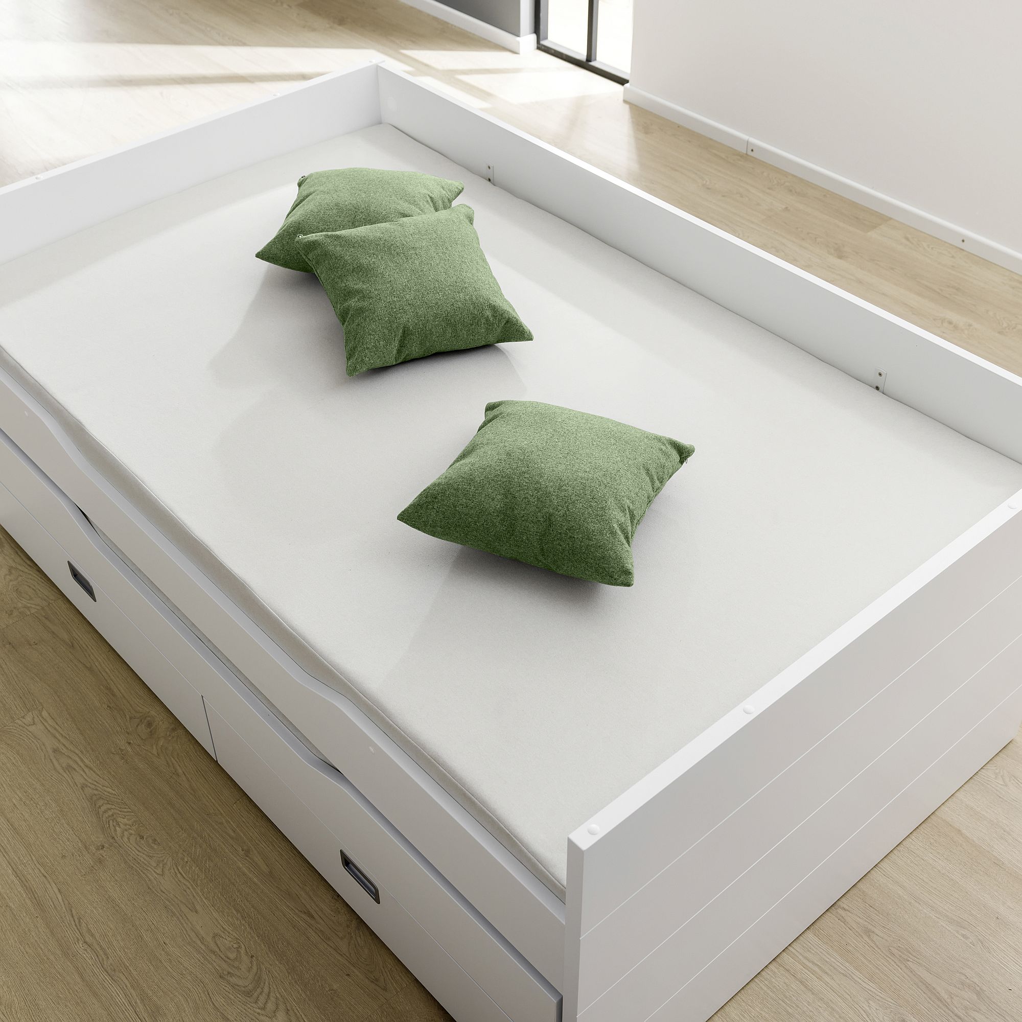Ein weißes Bett mit drei grünen Kissen, auf einem hellen Holzboden.