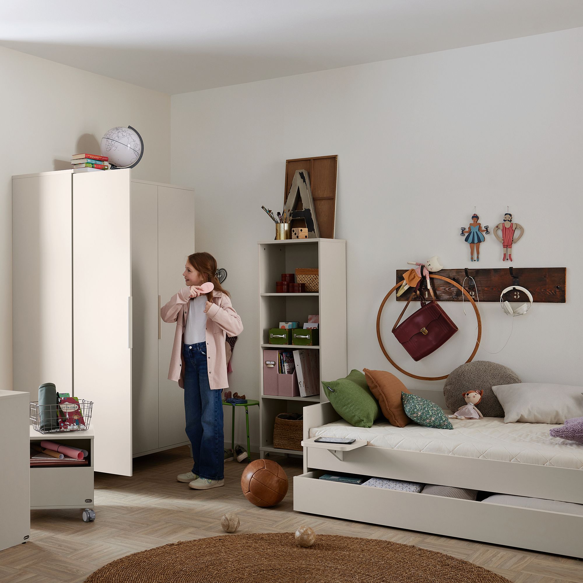 Ein Mädchen steht in einem aufgeräumten Schlafzimmer mit Bett, Kleiderschrank und Regalen.