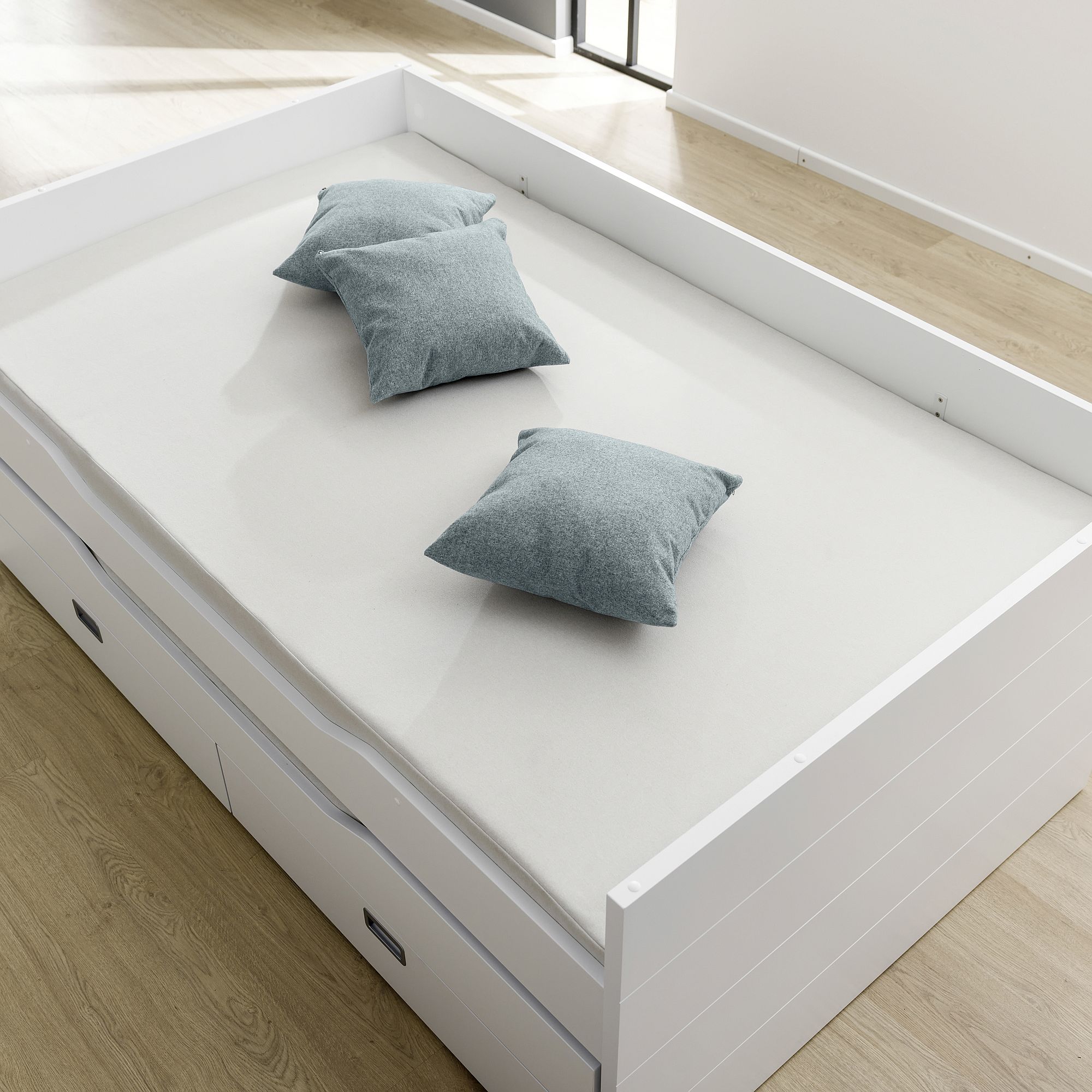 Ein weißes Bett mit drei grauen Kissen und Schubladen darunter, auf einem Holzboden.