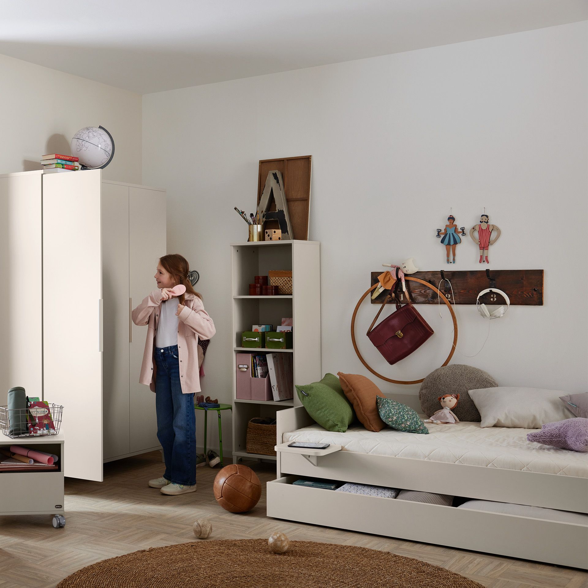 Ein Mädchen steht in einem aufgeräumten Schlafzimmer mit Bett, Kleiderschrank und verschiedenen Dekorationen.