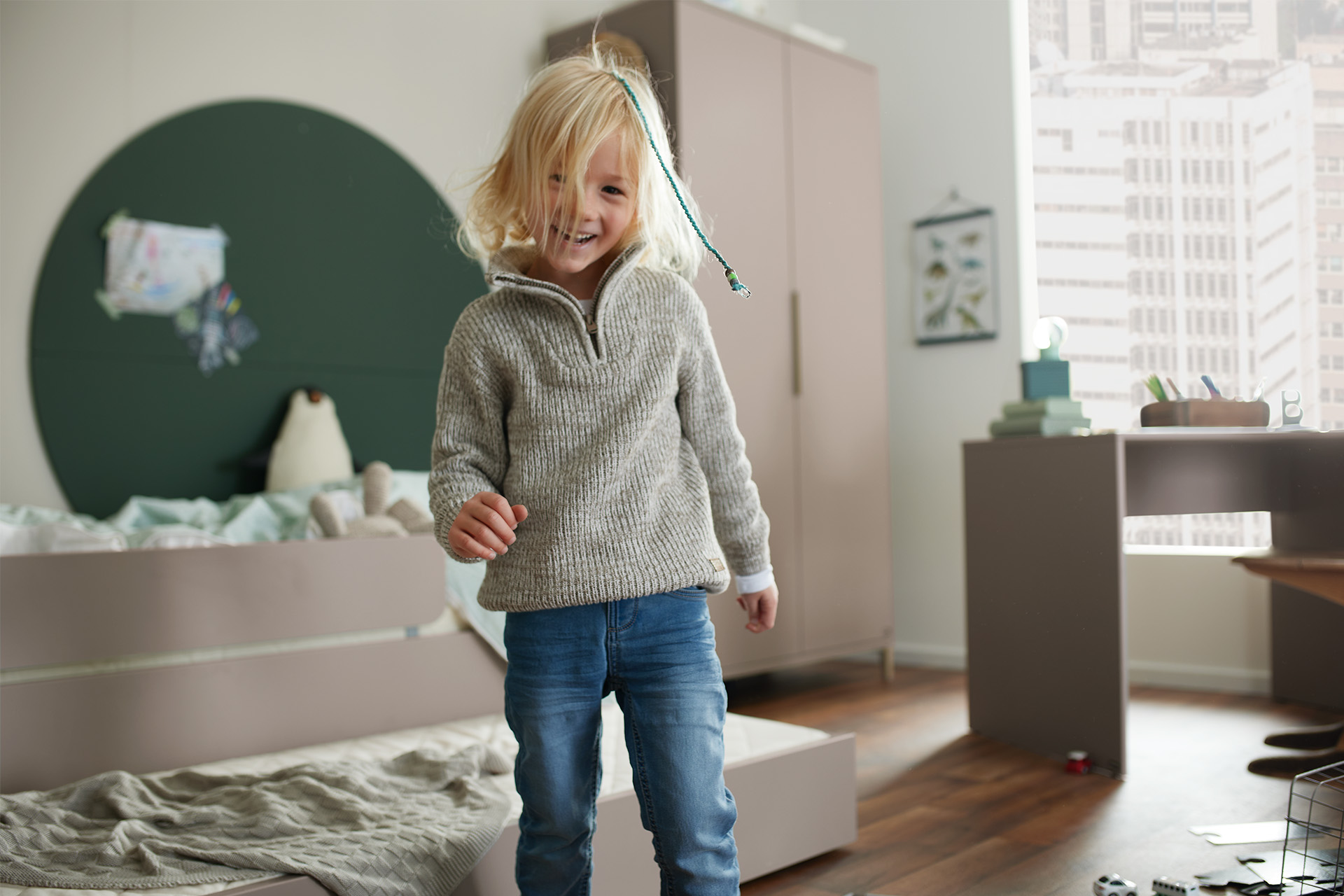 Ein lächelndes Kind mit blondem Haar steht in einem gemütlichen Schlafzimmer, trägt einen grauen Pullover und blaue Jeans.
