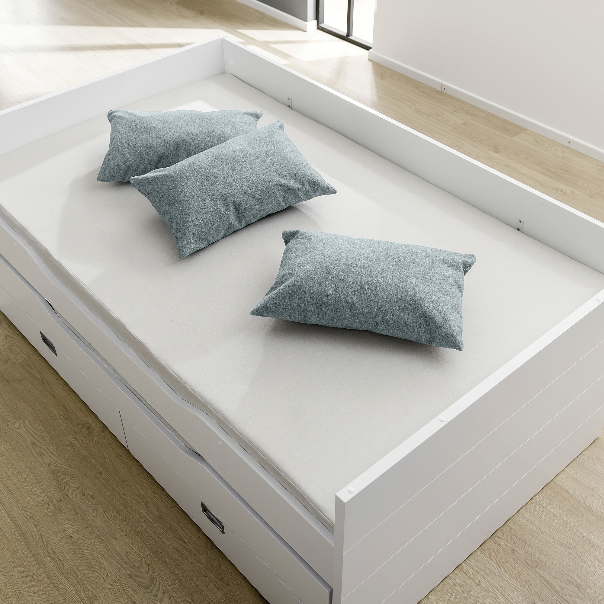 Ein weißes Bett mit drei grauen Kissen, in einem hellen Zimmer mit Holzfußboden.