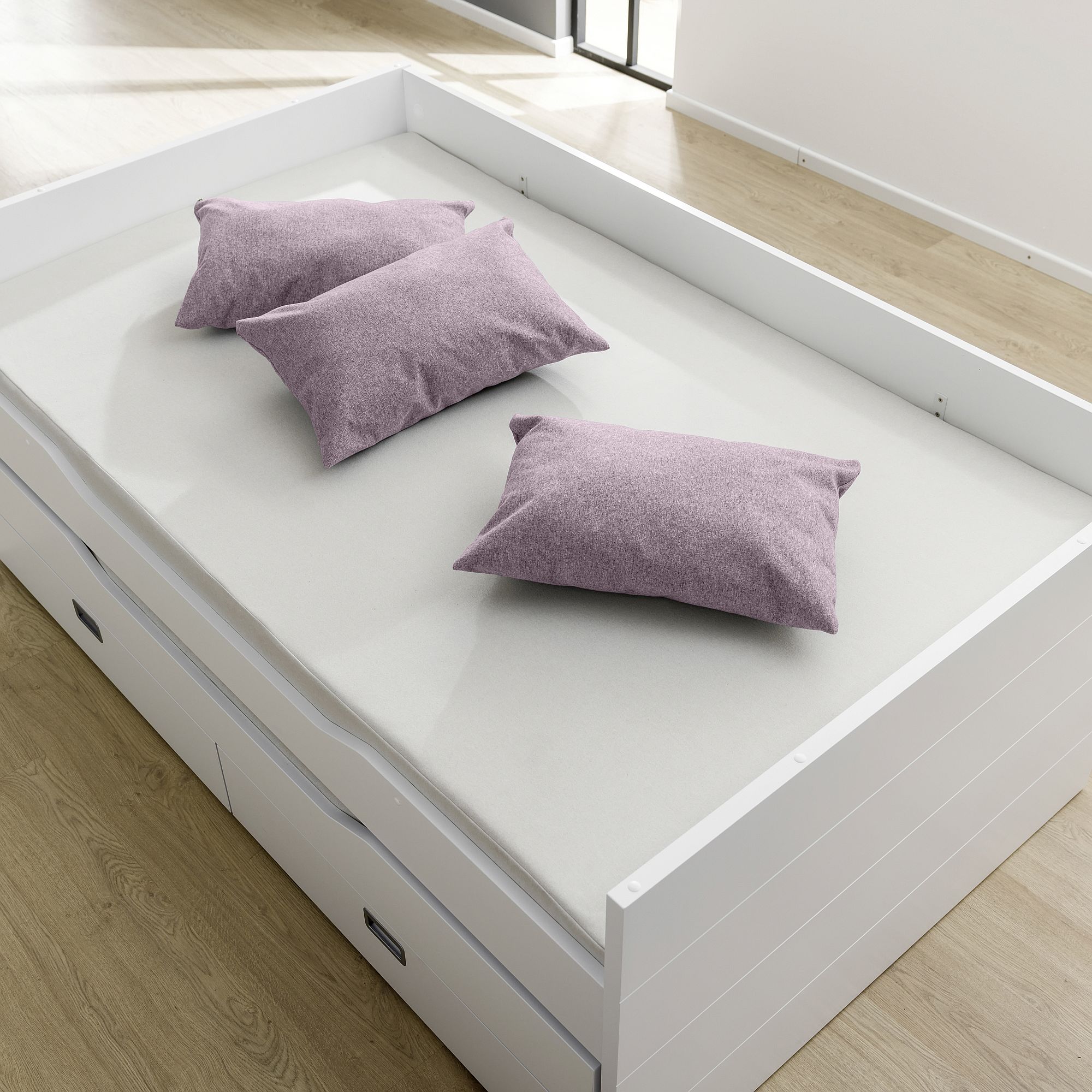 Ein weißes Bett mit drei lila Kissen auf einer hellen Matratze in einem hellen Raum.
