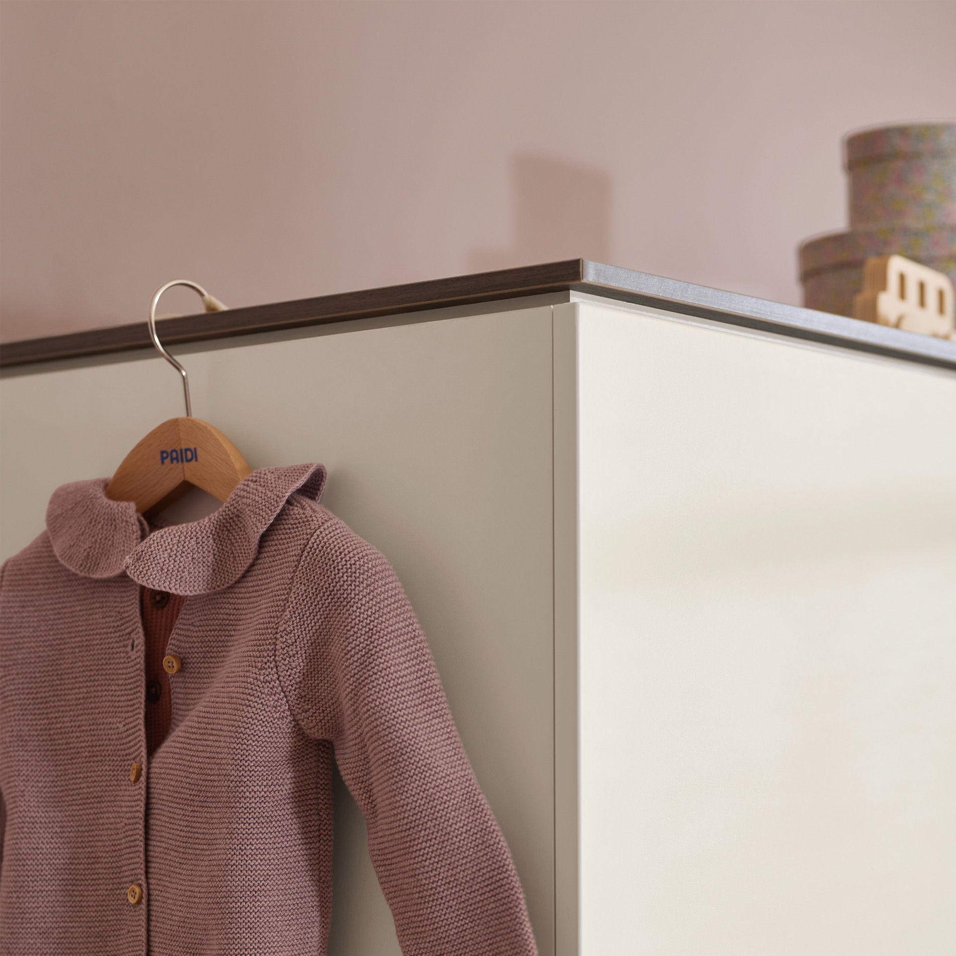 Rosa gestrickter Babypullover auf einem Bügel, der an der Ecke eines cremefarbenen Schranks hängt.