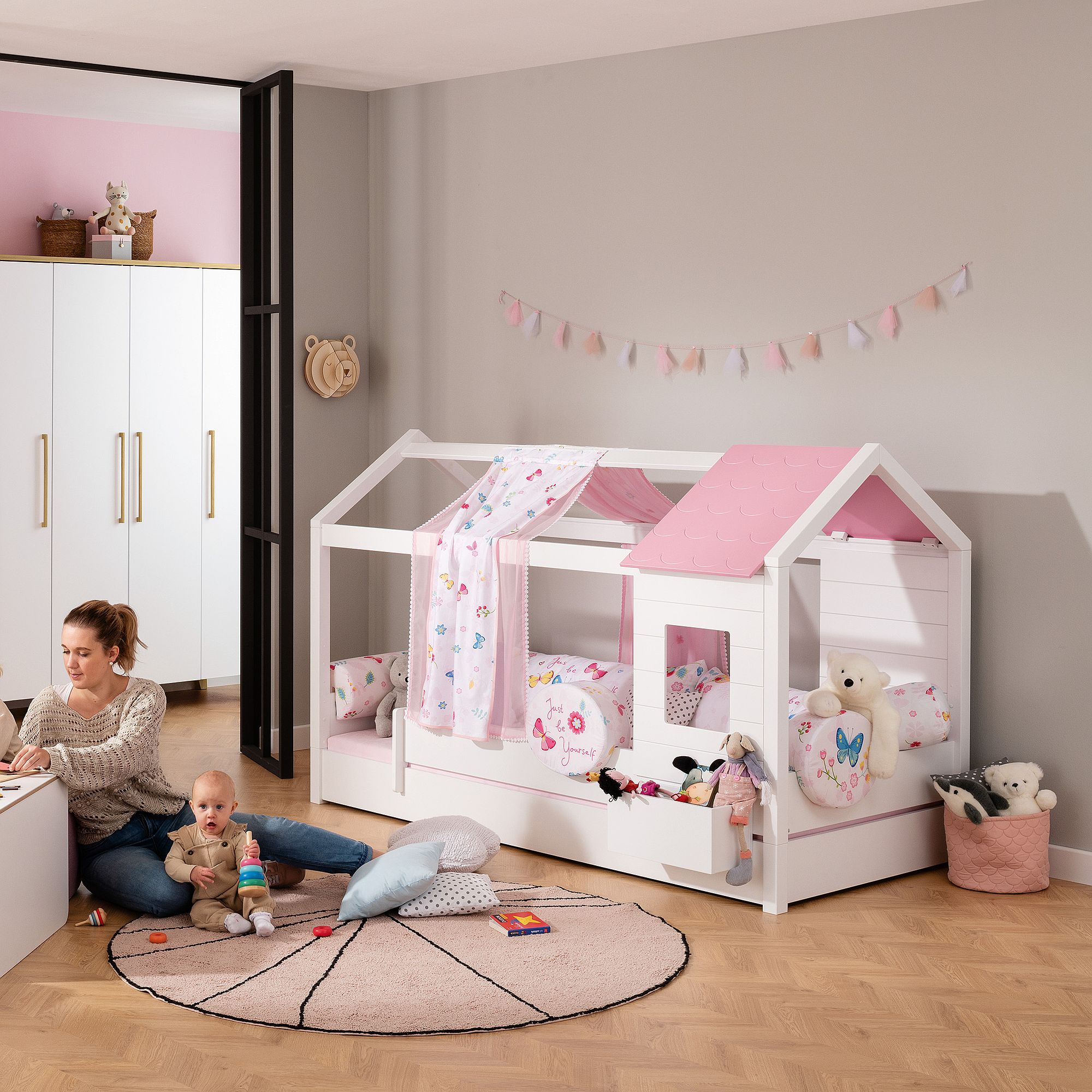 Frau und Baby spielen in einem pastellfarbenen Kinderzimmer mit einem rosa und weißen, hausförmigen Bett.