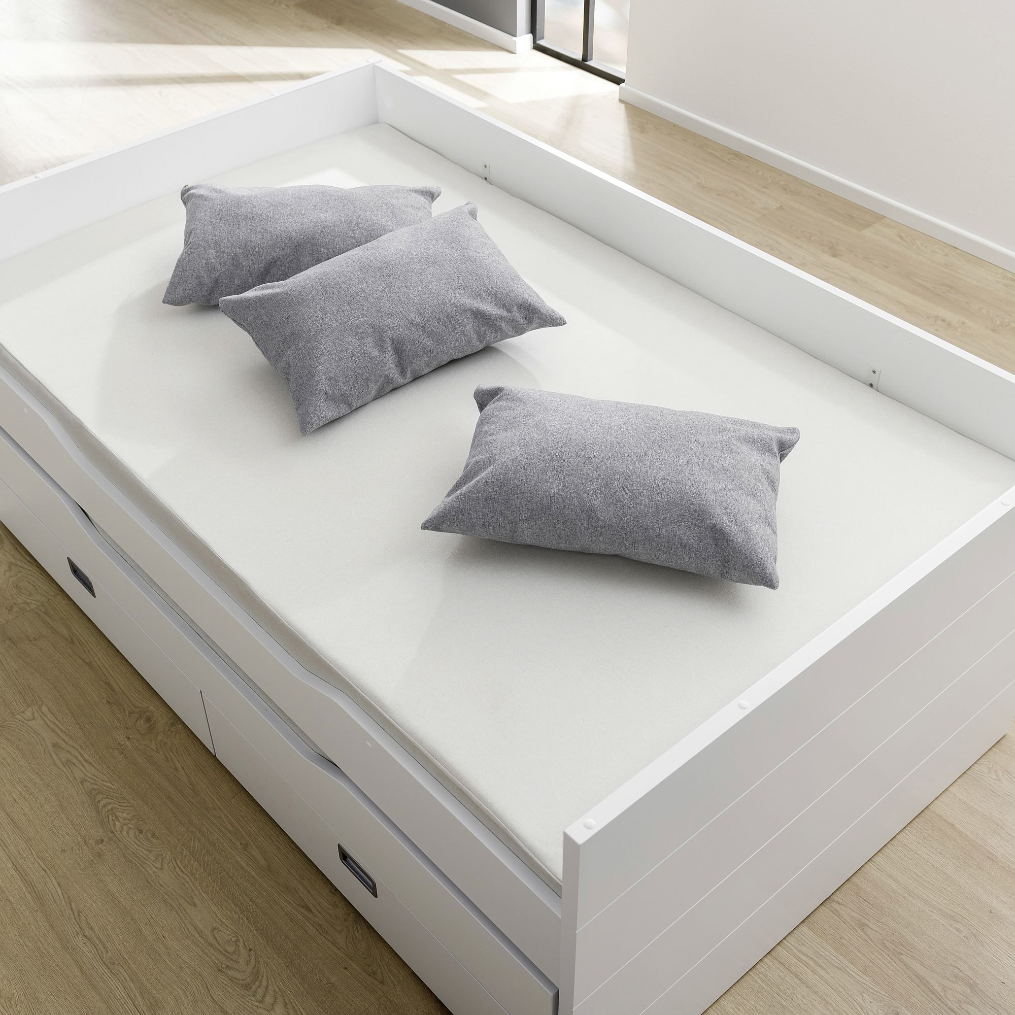 Ein weißes Bett mit drei grauen Kissen auf einem hellen Holzboden.