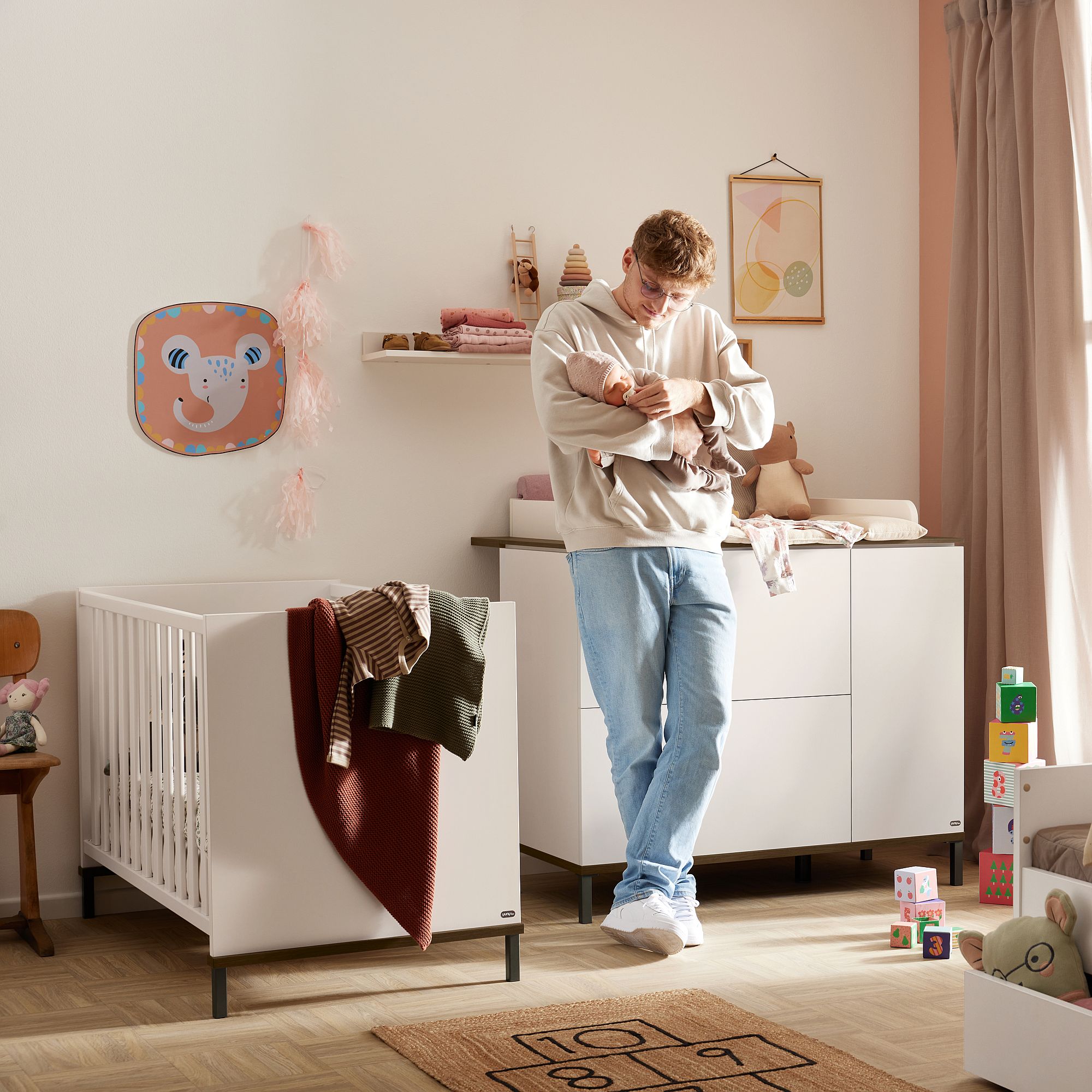 Ein Mann steht in einem Kinderzimmer und hält ein Baby neben einem Kinderbett und einem Wickeltisch.