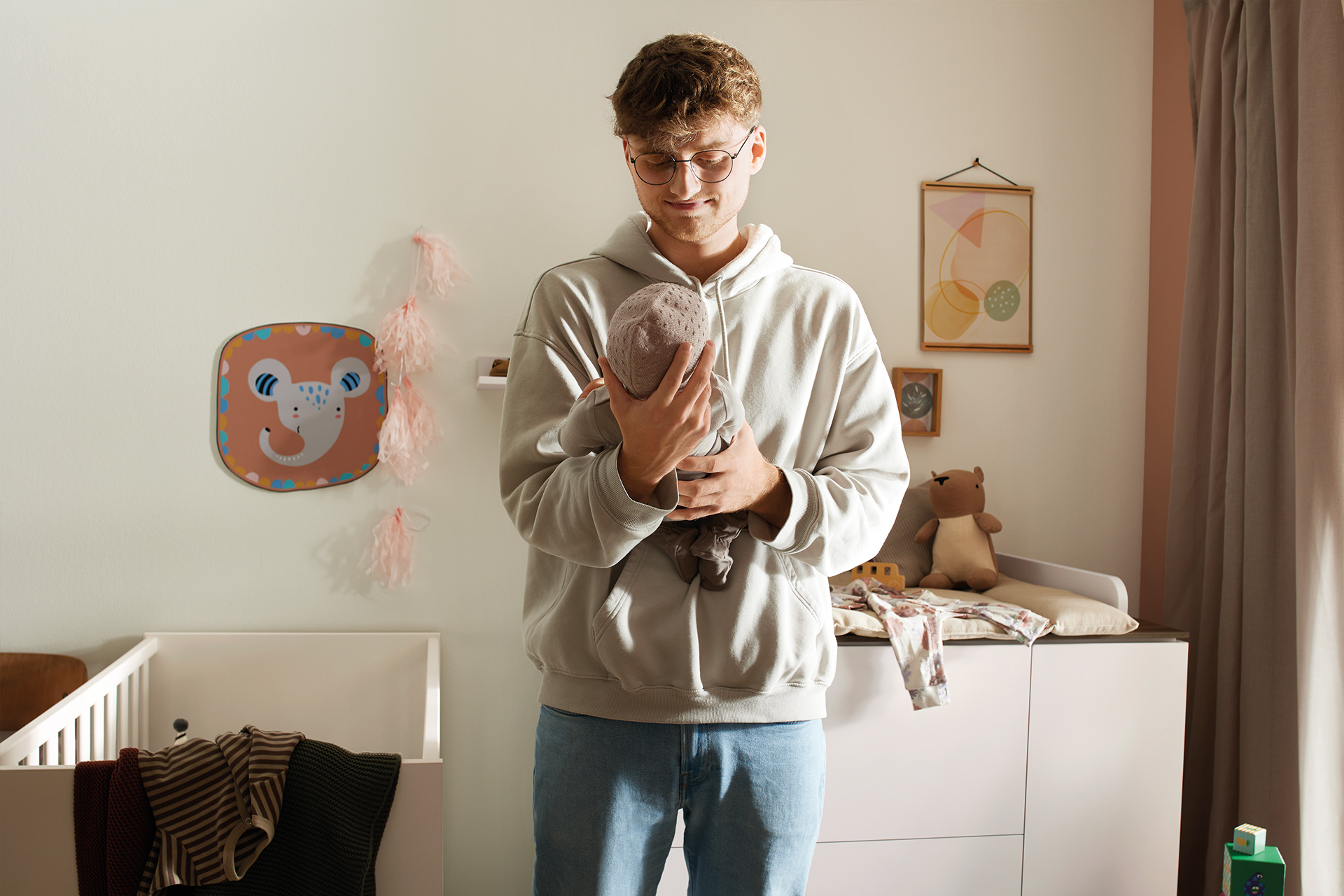 Ein Mann mit Brille hält sanft ein Baby in einem gemütlich eingerichteten Kinderzimmer.