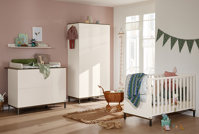 Modernes Kinderzimmer mit weißem Kinderbett, Kleiderschrank, Wickeltisch, Spielzeug, Weidenkorb und pastellfarbenem Dekor auf hellem Holzboden und rosa Akzentwand.