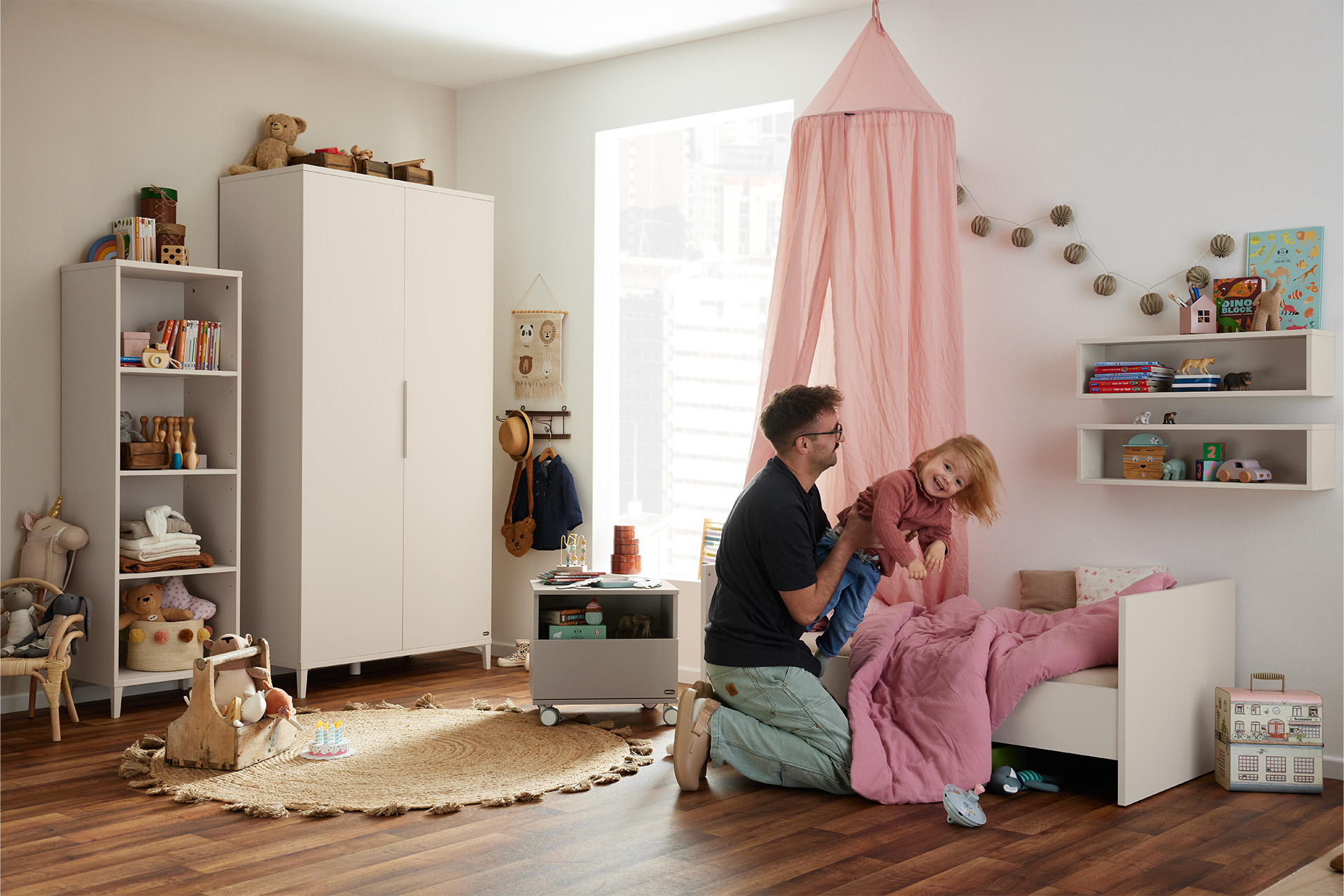 Ein Mann spielt mit einem kleinen Kind in einem gemütlichen, aufgeräumten Schlafzimmer mit Spielzeug, Regalen und einem rosa Baldachin über dem Bett.
