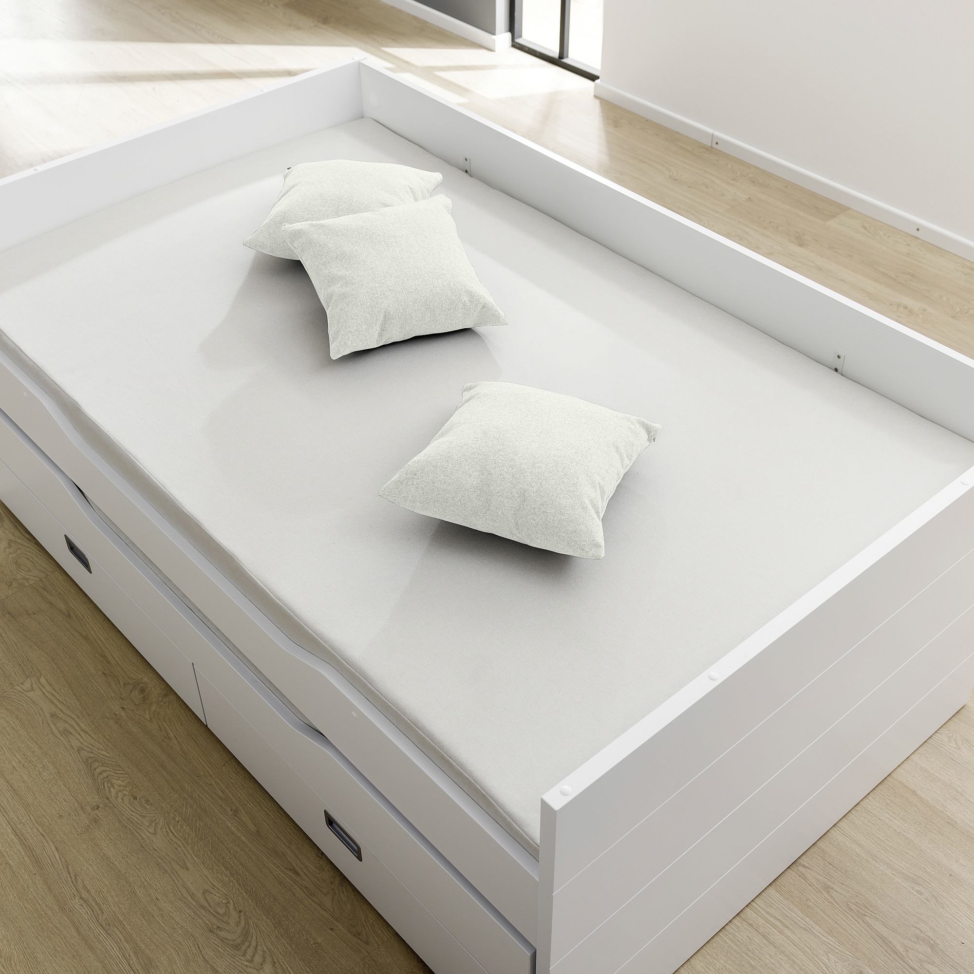 Ein weißes Bett mit drei weißen Kissen, auf einem hellen Holzboden.