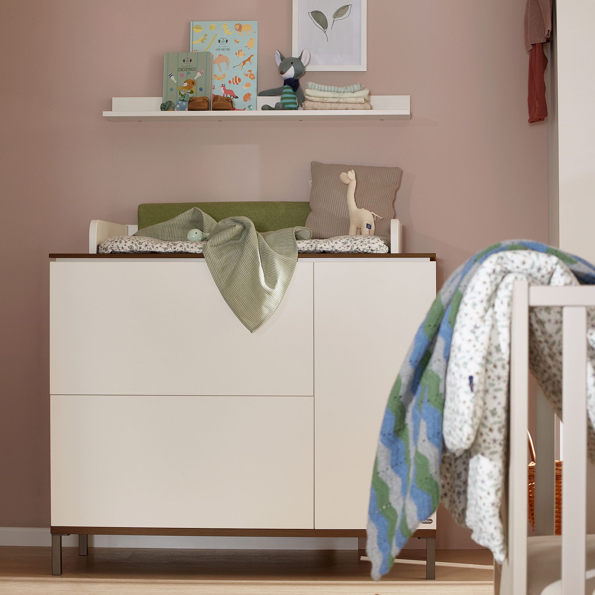 Modernes Kinderzimmer mit einem Wickeltisch, Spielzeug und Büchern auf einem Regal vor einer rosafarbenen Wand.