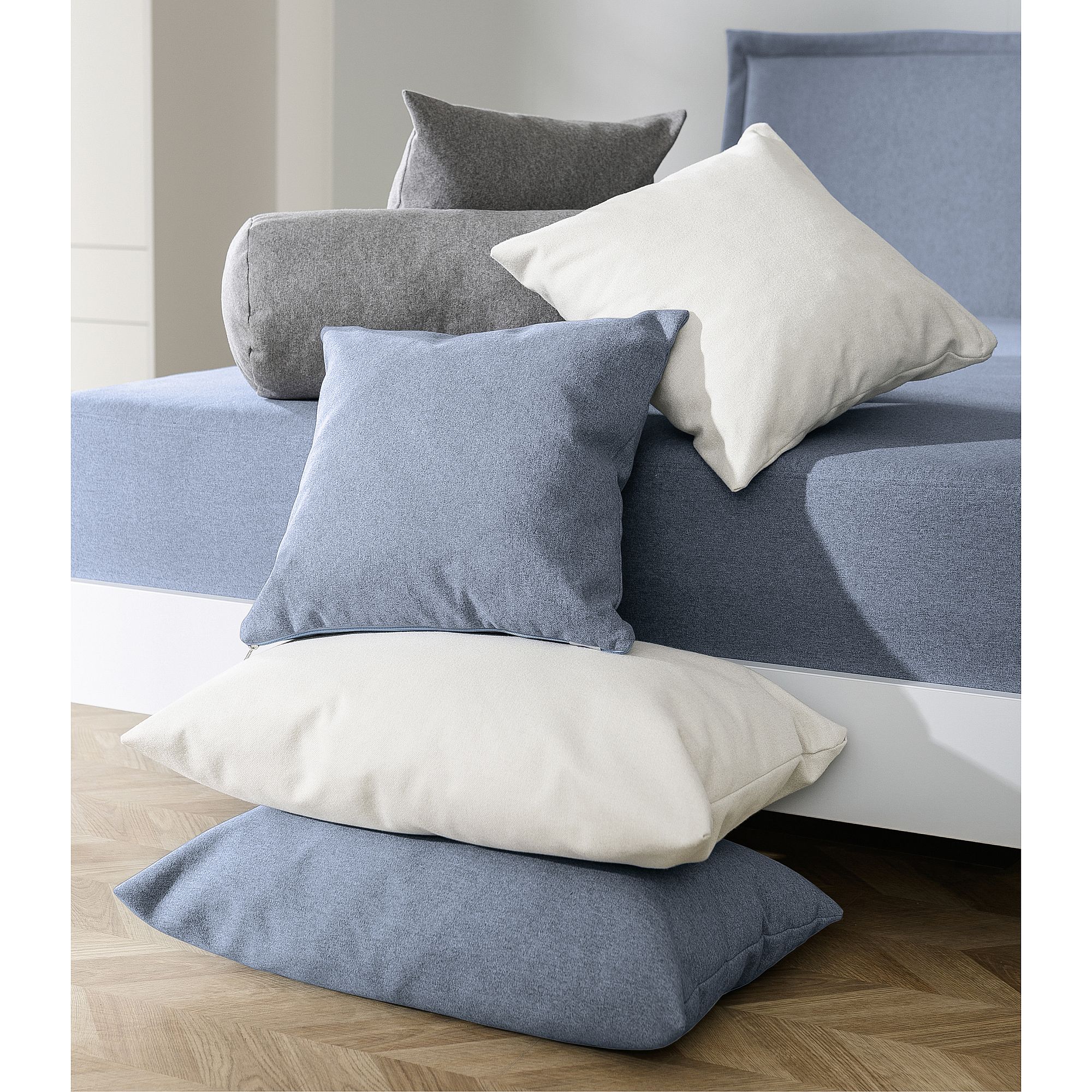 Ein Stapel blauer, grauer und weißer Kissen liegt auf und neben einem blauen Bett.