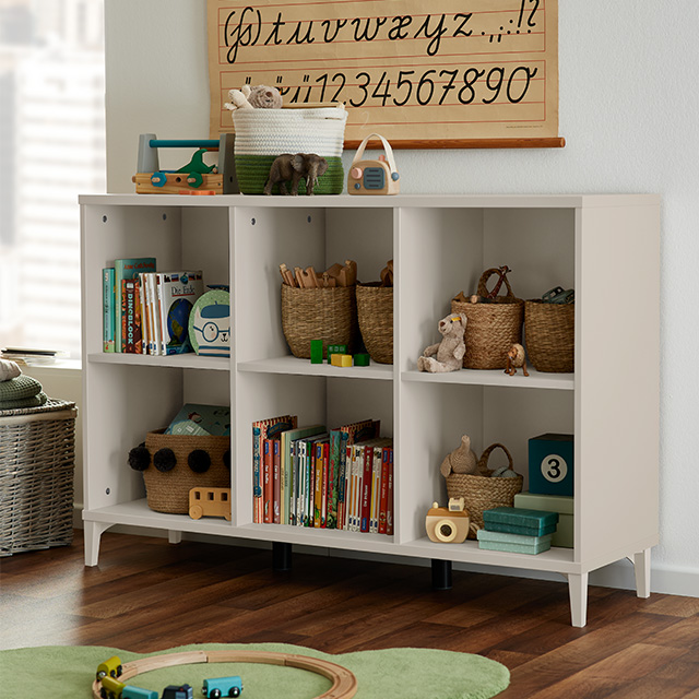 Ein weißes Regal mit Körben, Büchern und Spielzeug; an der Wand darüber hängen ein grüner Teppich und ein hölzernes Alphabetposter.